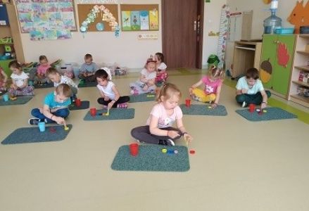 Dzieci siedzą przed dywanikiem, na nim mają kolorowe nakrętki i kubki