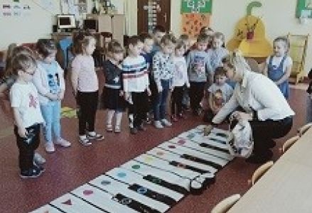 dzieci stoją przed matą do gry, nauczycielka rozdaje im kolorowe kształty