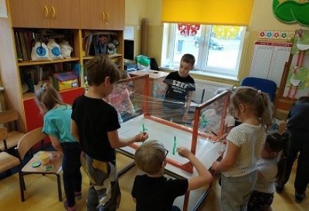 stolik obrócony do góry nogami dzieci stoją przy nim i malują pędzelkiem po foli, która otacza nogi stołu